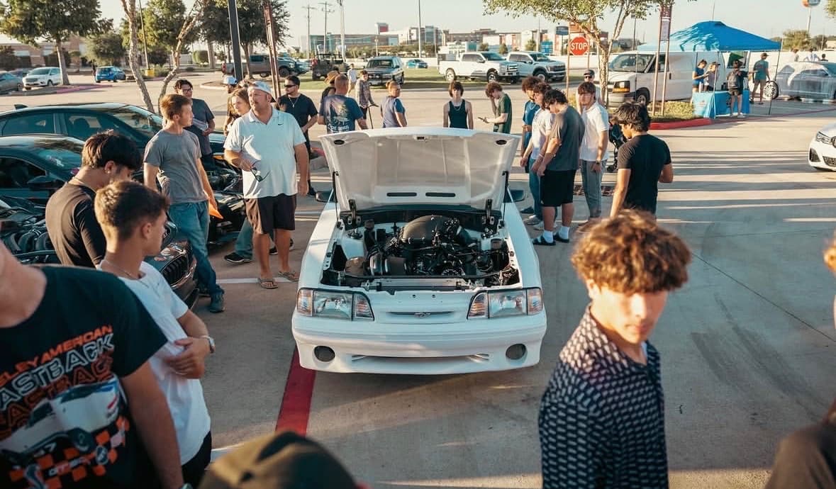 White Fox-body Mustang with the hood up drawing a crowd around the engine